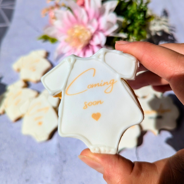 Pregnancy Announcement Biscuits: A close-up of a biscuit decorated with a baby onesie and the words "Coming Soon."