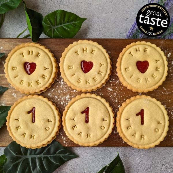 There are round biscuits on a wooden board, three with "The Biskery" and three with "1 Year Of."