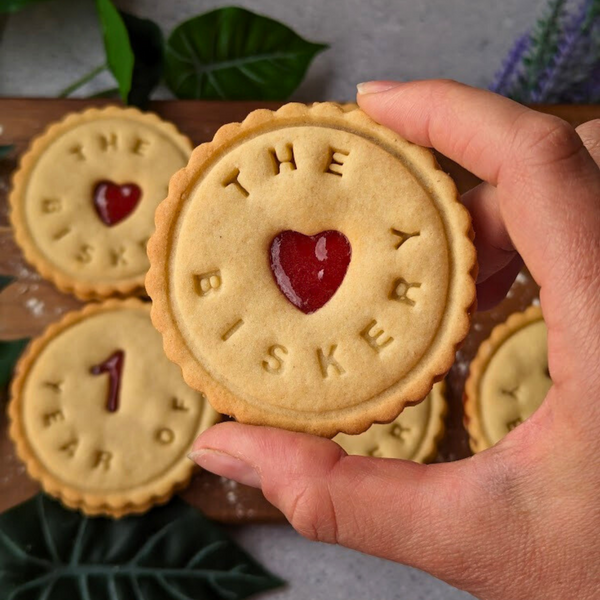 Hand holding a branded butter biscuit with "The Biskery" and "1 Year Of" messages celebrating a brand anniversary