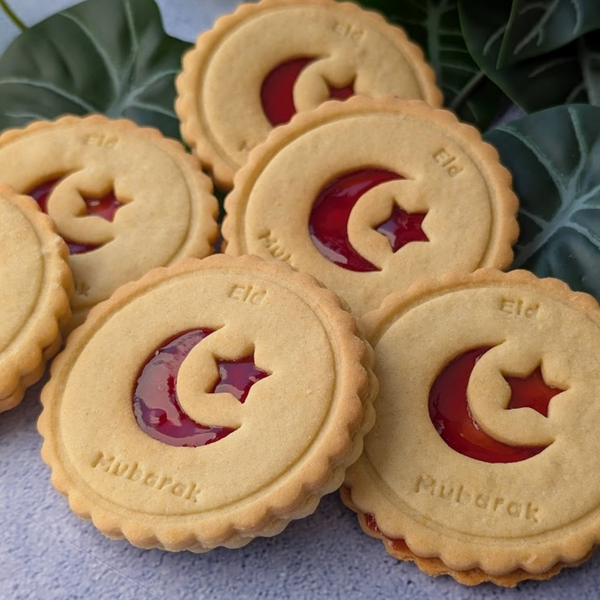 Golden Eid Mubarak Celebration Biscuits with red star and crescent jam fillings. Each biscuit is printed with "Eid Mubarak". Green leaves in background.