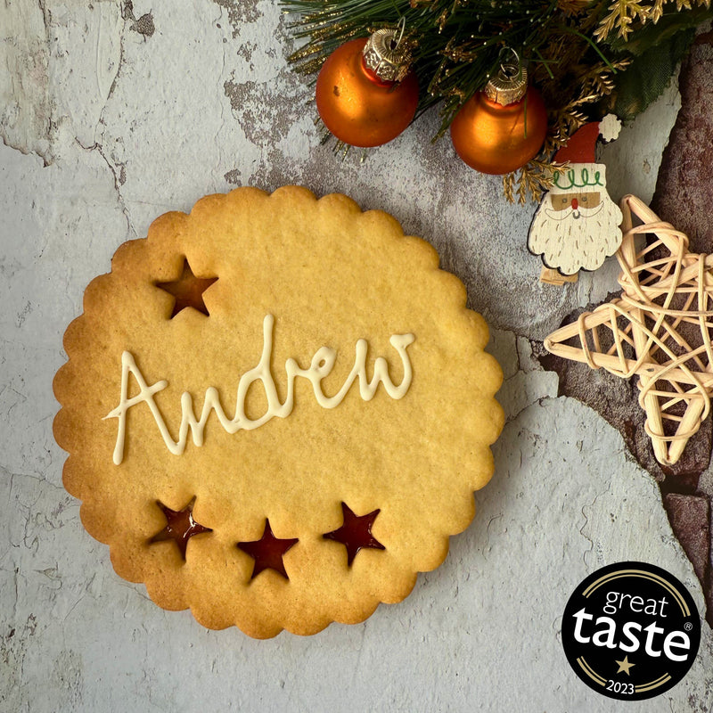 Giant Personalised Christmas Cookie in a gift box, decorated with white icing and the name "Andrew". Festive decorations and a Great Taste award logo are visible in the background.