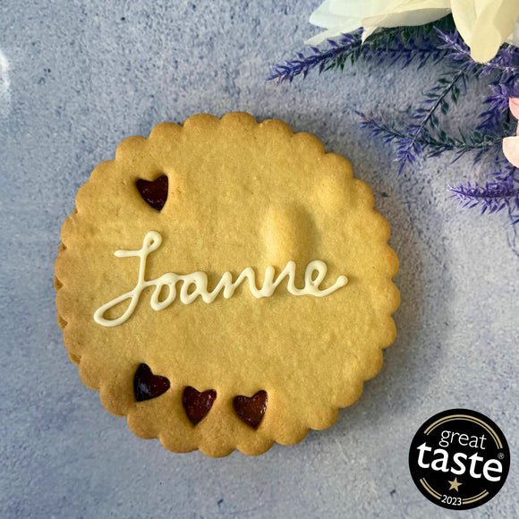 Giant Personalised Cookie in a gift box, decorated with white icing and the name "Joanne". Festive decorations and a Great Taste award logo are visible in the background.