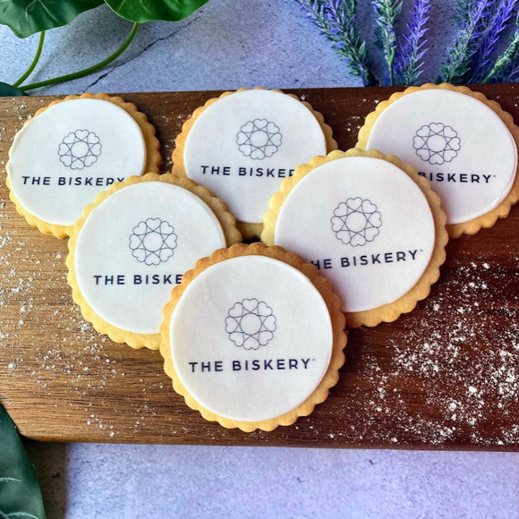 Six round branded cookies with The Biskery logo printed on white fondant icing, arranged on a wooden board. 