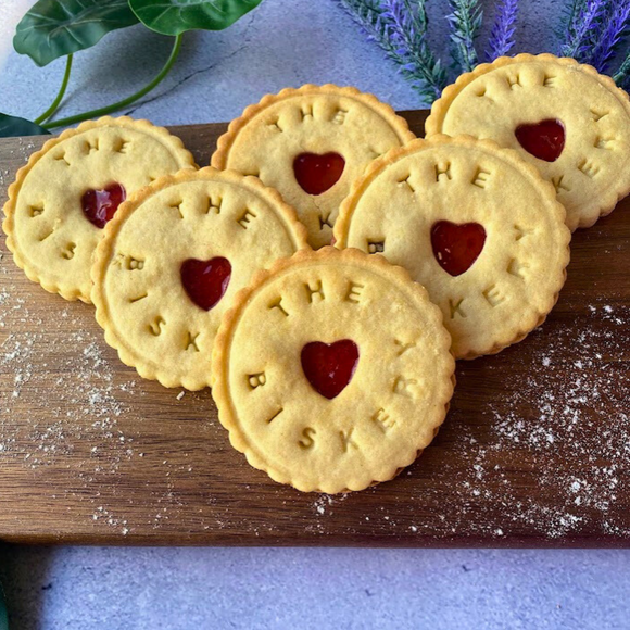  Six jam biscuits with "The Biskery" logo and heart-shaped cutouts arranged on a wooden board.