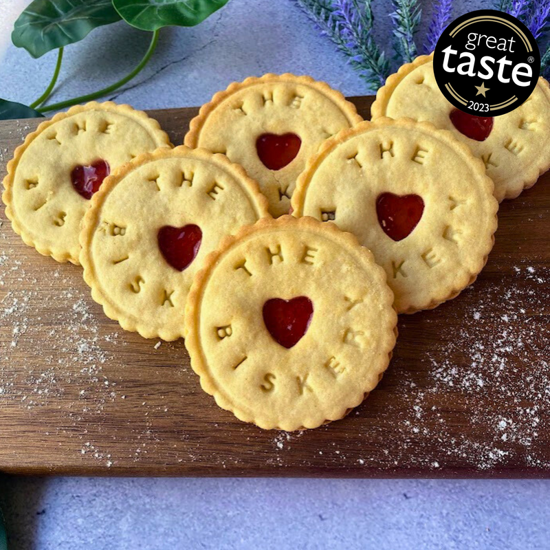  Six jam biscuits with "The Biskery" logo and heart-shaped cutouts arranged on a wooden board.