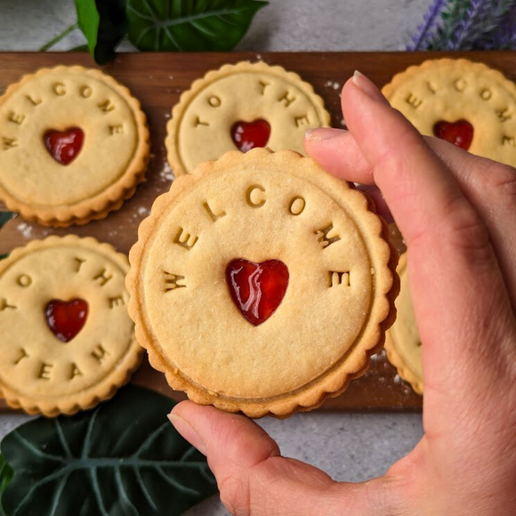 Hand holding a Great Taste award-winning jam cookies with a "Welcome" message & heart-shaped cutout. 