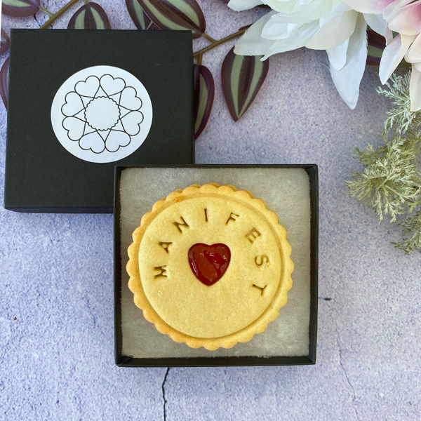 A round biscuit with a heart-shaped jam centre and the word "MANIFEST" embossed around it, presented in an open black gift box.