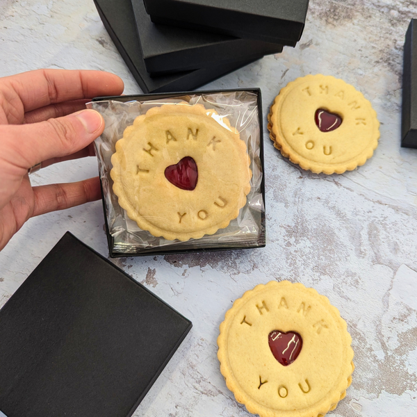 Several shortbread biscuits with "THANK YOU" message and heart-shaped jam filling, presented in black gift boxes.