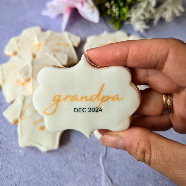 A hand holding a Pregnancy Announcement Biscuit shaped like a plaque, decorated with the word "Grandpa" and "Dec 2024" in gold lettering, with other biscuits in the background shaped like baby onesies.