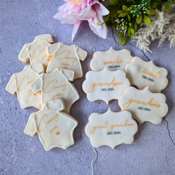 A collection of Pregnancy Announcement Cookies shaped like baby onesies and plaques, decorated with phrases like "Coming Soon" and "Grandma"