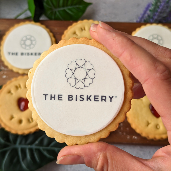 Hand holding a branded biscuit with The Biskery logo, from a gift box of 6 assorted treats. 