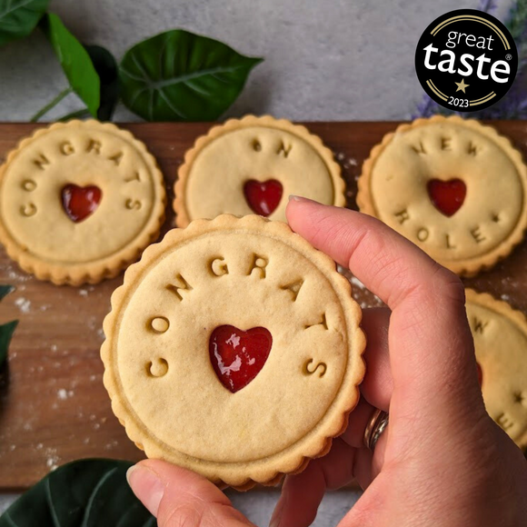 and holding a Great Taste award-winning jam biscuit with "Congrats" message and heart-shaped cutout.