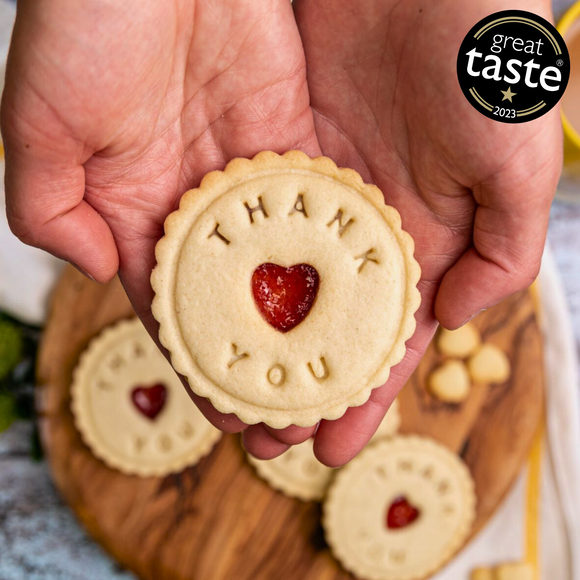 Hands holding a delicious, Great Taste Award-winning shortbread cookie with a heart-shaped jam centre and the words "THANK YOU" embossed on it. A perfect way to show appreciation with a sweet and thoughtful treat!