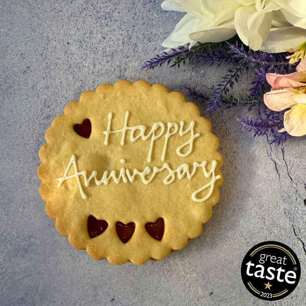 Giant Cookie | happy anniversary in a gift box, decorated with white icing and the name "Happy anniversary". Festive decorations and a Great Taste award logo are visible in the background.