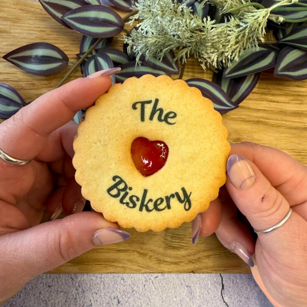 Hands hold a golden jam biscuit with a red heart centre. It's printed with "The Biskery". Wooden surface and greenery in the background. Personalised Printed Jam Biscuits.