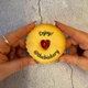 Hands hold a golden jam biscuit with a red heart centre. It's printed with "enjoy! The Biskery". Wooden surface and greenery in the background.