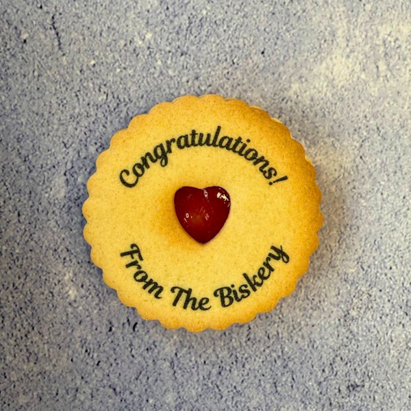 A close-up flat lay of a round, golden jam cookie with scalloped edges. It has a red heart-shaped jam centre and is printed with "Congratulations! From The Biskery". 