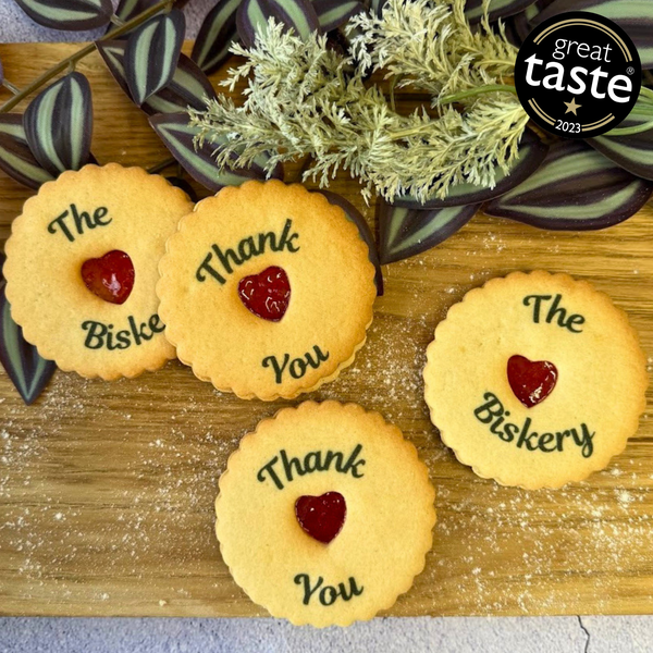 Four golden jam biscuits with red heart centres are arranged on a wooden surface with greenery. Each biscuit is printed with "The Biskery" or "Thank You". Great Taste 2023 logo visible.