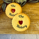 Two golden jam biscuits with red heart centres lie on a wooden surface. Both biscuits are printed with "The Biskery" in black lettering. 