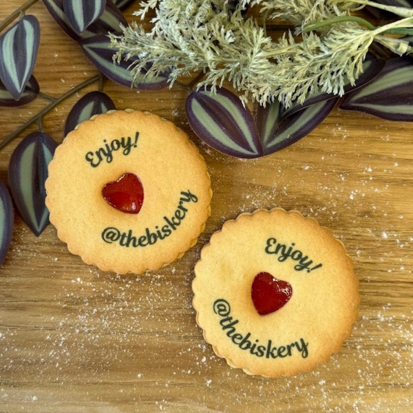 Two golden jam cookies with red heart centres are on a wooden surface with greenery. Each cookie is printed with "Enjoy!" and "@thebiskery". 