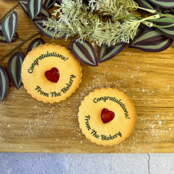 Two golden jam cookies with red heart centres are on a wooden surface with greenery. Each cookie is printed with "Congratulations" and "from the biskery"