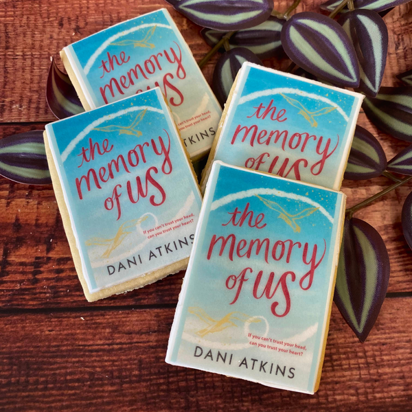 Four rectangular biscuits decorated to look like the book cover of "The Memory of Us" by Dani Atkins, arranged on a wooden surface with a leafy green plant.